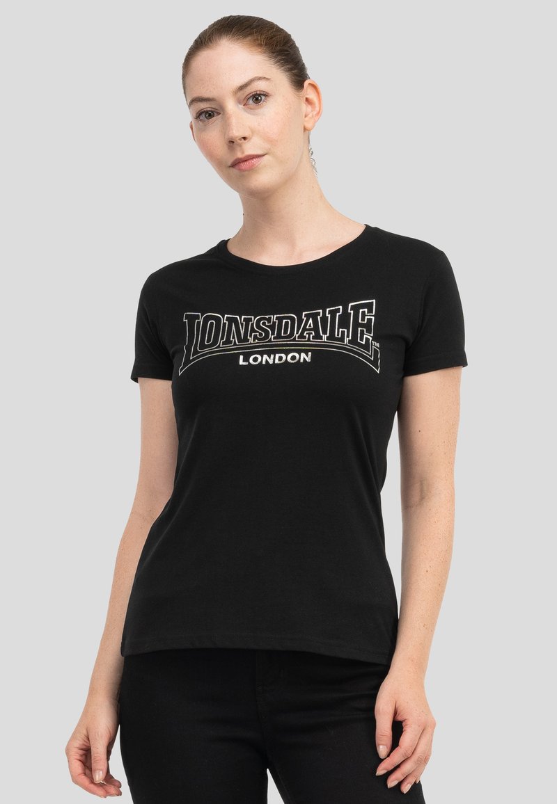 Woman with hair tied back wearing a black Lonsdale London t-shirt and black pants against a plain gray background.