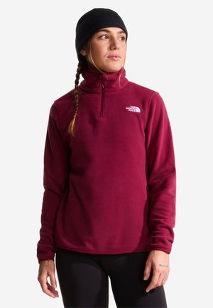 Vrouw met een zwarte muts en een bordeauxrood fleecejack met rits en The North Face-logo, kijkt iets naar rechts, draagt een zwarte broek.