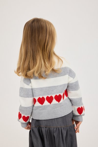 Enfant aux cheveux blonds mi-longs portant un pull rayé gris et blanc avec des cœurs rouges, debout de dos sur un fond uni.