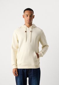 Cremfarbener Hoodie mit einer strukturierten Oberfläche, Fronttasche und verstellbarem Kordelzug mit braunen Akzenten. Getragen mit dunkelblauen Jeans.