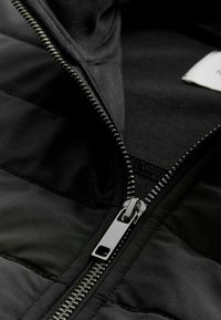 Schwarze, mit Reißverschluss versehene Jacke aus gestepptem Stoff mit glatter Innenauskleidung. Sichtbare silberne Reißverschlussbeschläge und Nähdetails.