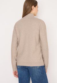 Beige, gerippter Strickpullover mit hohem Kragen und lockerer Passform, ausgestattet mit langen Ärmeln und einem Saum, der auf der Hüfte sitzt, kombiniert mit blauen Jeans.