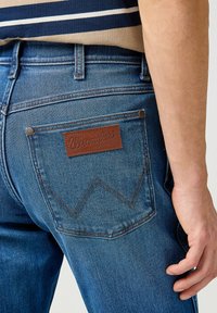 Vaqueros de mezclilla azul para hombre con un parche de cuero con el logotipo, costuras zigzagueantes en el bolsillo trasero y remaches de metal en los laterales.