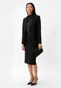 Schwarze maßgeschneiderte Blazer mit einem einzigen Knopf, kombiniert mit einem glitzernden Rollkragenpullover und einem schwarzen Bleistiftrock. Accessoires sind eine schwarze Clutch und spitze Absätze.