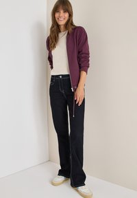 Giacca bomber bordeaux con zip, maglione beige a maglia, jeans neri a campana con cuciture bianche e sneaker bianche con accenti blu.