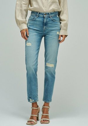 Mujer con jeans desgastados de mezclilla azul claro, blusa beige de manga larga y sandalias beige de tacón alto, posando sobre un fondo liso.