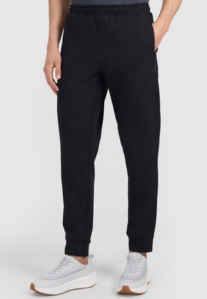 Hombre con pantalones deportivos negros y zapatillas blancas, de pie con una mano en el bolsillo frente a un fondo sencillo.