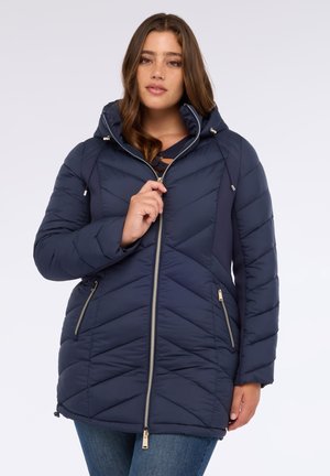 Giacca puffer blu navy con cappuccio, caratterizzata da cuciture a zigzag, chiusura zip frontale e tasche laterali. Texture morbida e trapuntata. Accenti dorati visibili.