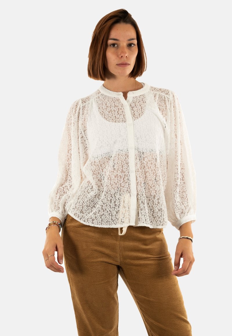 Blusa bianca di pizzo con maniche a sbuffo, scollo rotondo e chiusura frontale con bottoni; tessuto trasparente su un top solido; indossata con pantaloni di velluto a coste marroni.