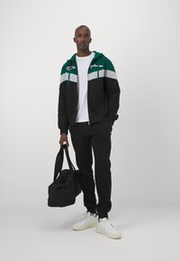 Sudadera con cremallera negra y azul verdoso con detalles grises, combinada con pantalones de chándal negros y zapatillas blancas. El modelo sostiene una bolsa de deporte negra.