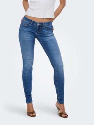 Kvinde iført mellemhøje slim blå jeans og brune åbne hælede sandaler, stående med hænderne bag ryggen.