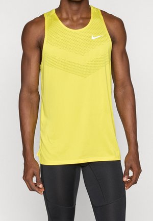 Muscular person wearing a sleeveless yellow Nike sports top and tight black athletic leggings standing with arms relaxed.