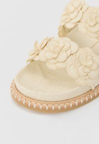 Beige sandāles ar ziedu rotājumiem, austo augšpusi, platformas zoli un šūtu ādas apdari. Tekstūrēta pēdu gultne labākai saķerei.