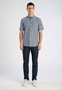 Camicetta a maniche corte con bottoni in tessuto intrecciato blu-grigio, abbinata a jeans scuri e sneakers bianche. Design semplice con colletto e bottoni frontali.