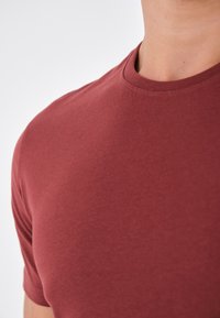 Rotes Baumwoll-T-Shirt mit Rundhalsausschnitt und kurzen Ärmeln. Der Stoff wirkt glatt mit einem leichten Glanz. Saubere Nähte und keine sichtbaren Muster.