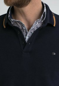 Polo shirt de couleur marine avec une finition texturée, présentant un imprimé floral à l'intérieur du col et un accent moutarde contrastant sur le bord du col. Détail de fermeture à boutons.