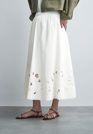 Frau trägt einen weißen Midirock mit floralem Ausschnitt-Saum, braune flache Sandalen und eine olivgrüne cropped Jacke, steht auf grauem Boden.