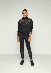 Sweatshirt preto com detalhes em dourado e padrão de riscas de tigre; calças de fato pretas combinando com um design semelhante; usadas com ténis pretos.