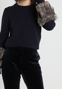 Maglione blu navy con dettagli in pelliccia sintetica grigia a trama ai polsini, abbinato a pantaloni di velluto neri, evidenziando un design elegante e aderente.