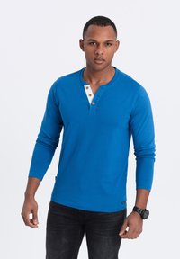 Lange mouwen blauwe henley-shirt van katoen met witte knoopsluiting en bruine knopen; gedragen met donkere jeans en een zwarte horloge.