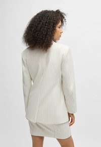 Valkoinen pinstripes-takki, jossa on rakenteelliset hartiat ja pidentynyt siluetti, yhdistettynä vastaavaan pinstripes-minihameeseen.