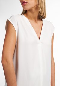 Blusa bianca con scollo a V, maniche a cappuccio e un delicato dettaglio con cucitura centrale. Tessuto liscio e leggero con vestibilità rilassata.