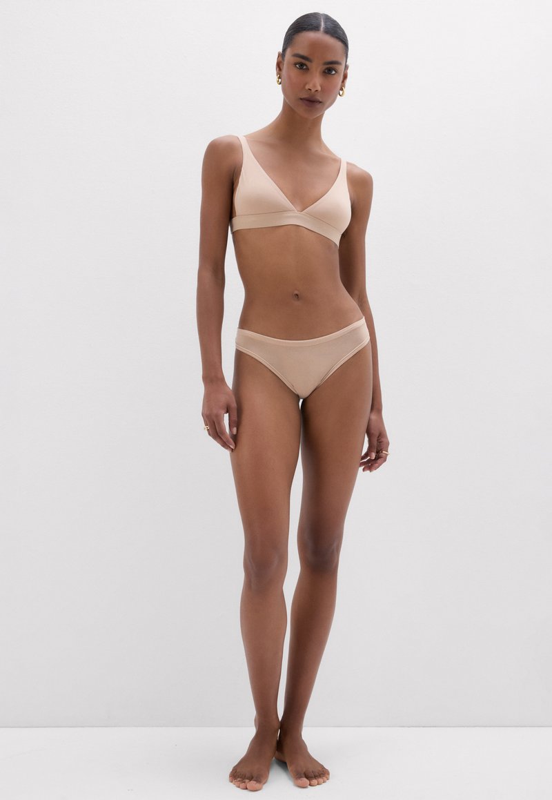 Woman with dark hair in a beige bra and underwear set standing barefoot against a plain white background.