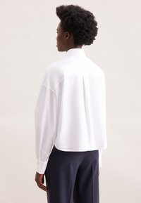 Camicia bianca oversize con orlo curvo, maniche lunghe e polsini con bottoni, caratterizzata da una piega sul retro. Tessuto con texture liscia.