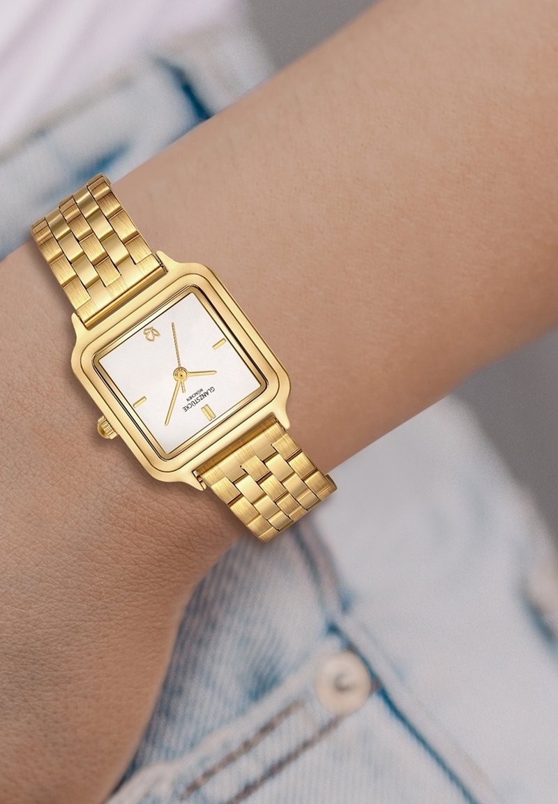 Kuldkattega ristkülikukujuline kell valge näo, peente tunniindikaatorite ja punutud metallrihmaga. Kell on randmel heleda denimkangaga.