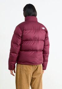 Burgundy puffjacka med hög krage, kviltad design och lätt glans. Har North Face-logotypen på övre ryggen.