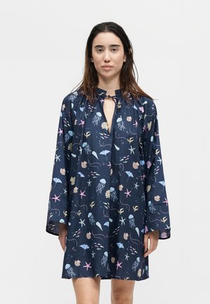 Γυναίκα που φοράει ένα χαλαρό μπλε φόρεμα με ροζ, μπλε και μπεζ αποτυπώματα θαλάσσιων πλασμάτων και κοχυλιών, στέκεται μπροστά σε απλό φωτεινό φόντο.