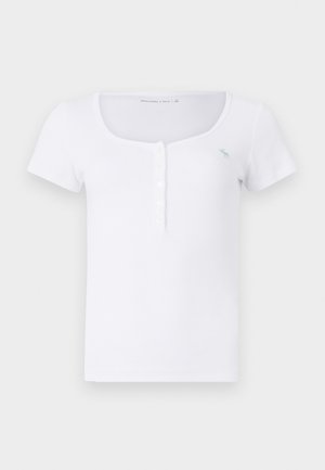 Hvit kortermet t-skjorte laget av ribbestrikket stoff, med scoop-hals og knappelukking med en liten blå logo på brystet.