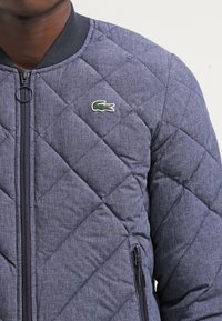 Překládaná bomber bunda v námořnické modři s texturovaným povrchem, vybavená zipovým zapínáním, ribbed límcem a vyšívaným zeleným logem na hrudi.