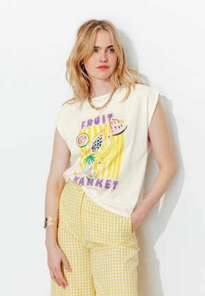 Femme portant un top blanc sans manches avec un imprimé de fruits colorés et un pantalon vichy jaune, accessoirisée avec un collier et un bracelet en or.