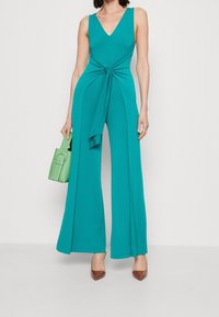 Mono verde azulado con escote en V, detalle de cintura anudada y piernas anchas. Combinado con un bolso verde y tacones marrones de punta. Tejido suave.