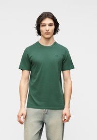 Νεαρός άνδρας με μεσαίου μήκους σκούρα μαλλιά, φορώντας απλό πράσινο κοντομάνικο μπλουζάκι και ανοιχτόχρωμα μπλε τζιν, στέκεται μπροστά σε λευκό φόντο.