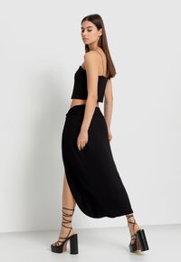 Schwarzes, kurzes Tanktop, kombiniert mit einem schwarzen Maxirock mit hohem Schlitz. Das Model trägt schwarze, geschnürte Plateausandalen.