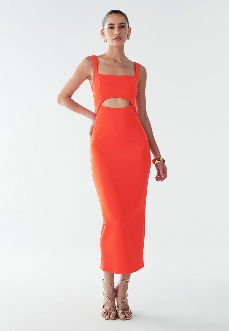 Vrouw in een aansluitende, mouwloze oranje midi-jurk met een vierkante halslijn en een uitsnijding bij de buik, gecombineerd met gouden strappy hoge hakken.