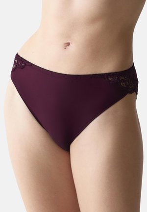 Kobieta ubrana w ciemnofioletowy, bikini-styl bieliznę z koronkowymi detalami po bokach na jednolitym jasnym tle.