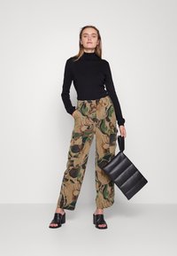 Camisola de gola alta preta, calças cargo com padrão de camuflagem e sandálias de salto bloco pretas. Segurando uma mala de ombro preta acolchoada.