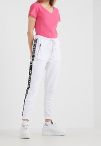 Camiseta de cuello en V rosa combinada con pantalones de chándal blancos que presentan una franaja lateral negra con el texto "TRUE RELIGION" y bolsillos con cremallera. Zapatillas blancas.