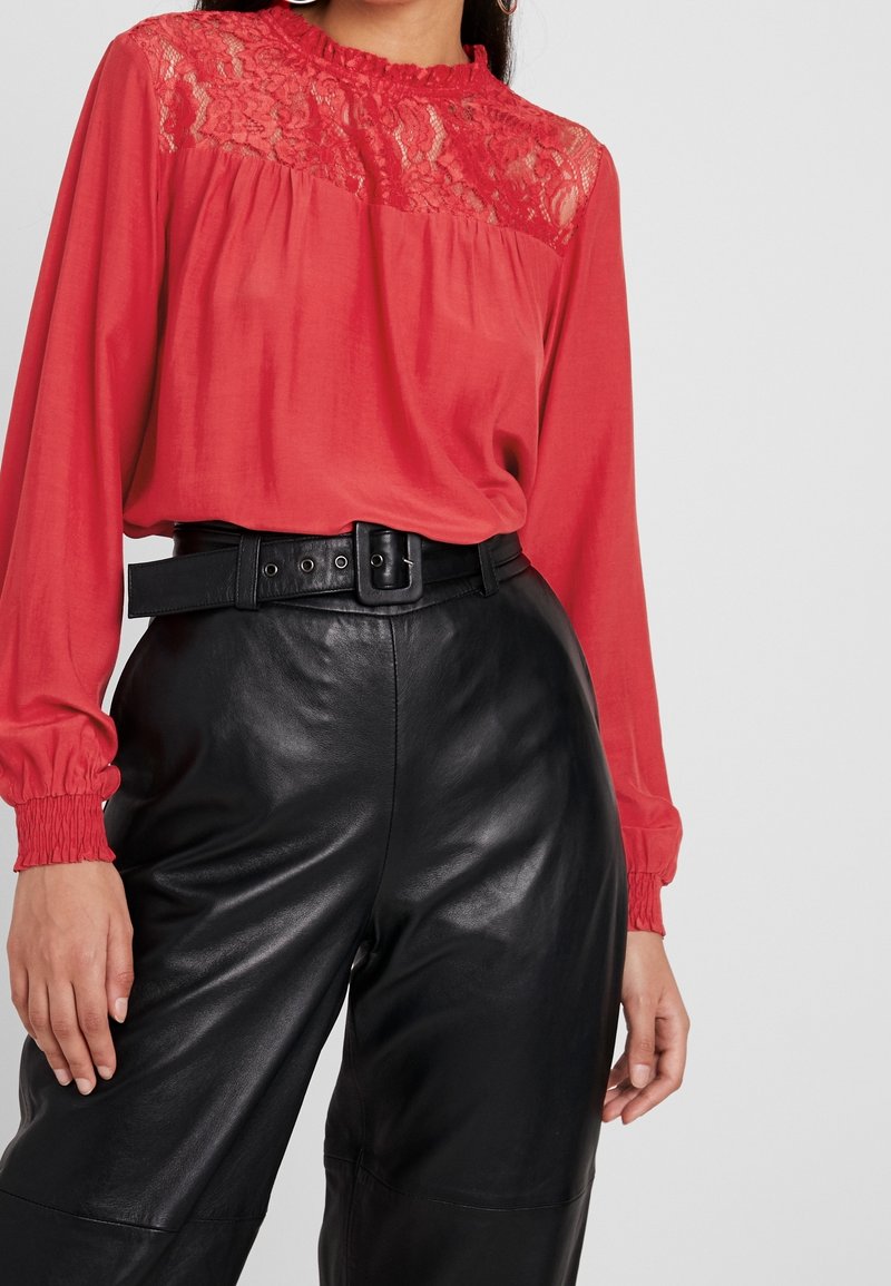 Femme portant un chemisier rouge à manches longues avec empiècement en dentelle et un pantalon en cuir noir taille haute avec ceinture assortie.