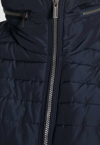 Marinblå puffjacka med quiltat mönster, vertikal sömnad och en central dragkedja med metallpull. Inkluderar två sidofickor med dragkedja.