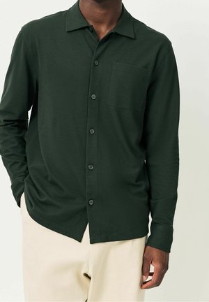 Homme portant une chemise boutonnée à manches longues vert foncé avec une poche poitrine et un pantalon beige clair sur un fond uni.
