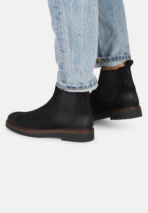 Botas Chelsea de ante negro con laterales elásticos, puntera redonda y suela plana de goma, combinadas con jeans de tiro bajo en azul claro con el dobladillo enrollado.