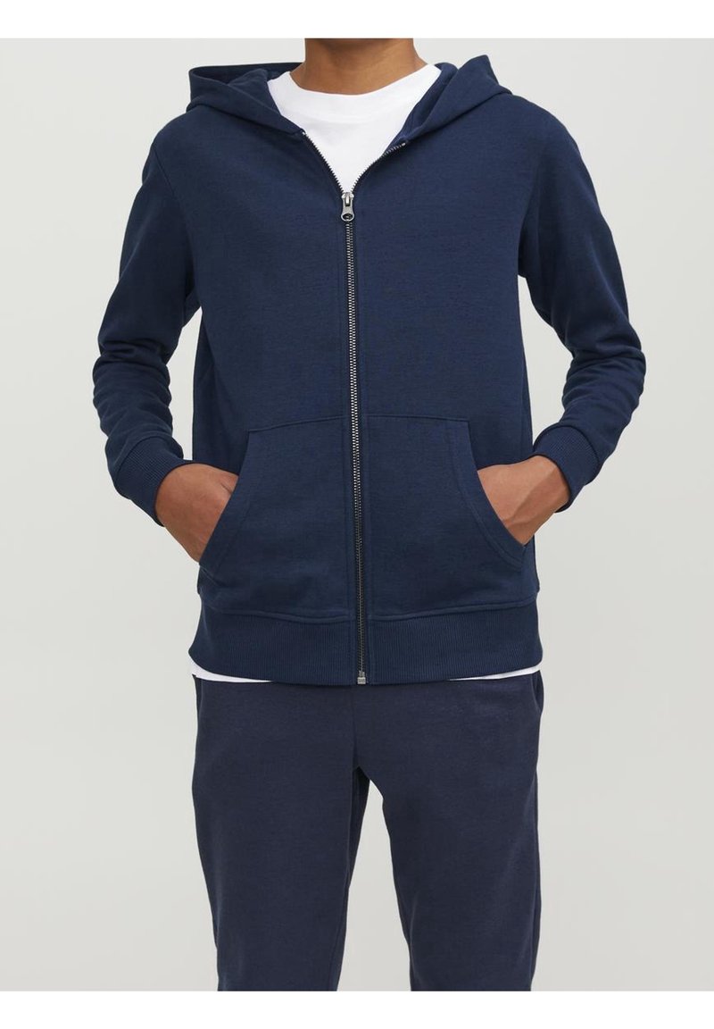 Sweat à capuche zippé marine en tissu doux, avec une poche frontale et une capuche à cordon. Poignets et ourlet côtelés, porté sur une chemise blanche.