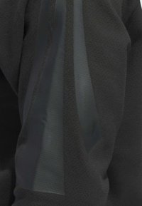 Schwarzes Sportoberteil mit strukturiertem Stoff und glatten, durchsichtigen Akzenten entlang der Schultern und Seitennähte.