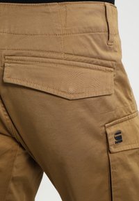 Tan cargo-byxor har flera fickor, ett slitstarkt tyg och kontrasterande sömmar. En logotypmärke syns på vänster ficka.