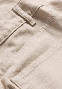 Beige cotton fabric with a smooth texture and visible stitching. Features a narrow strap and pocket detail.