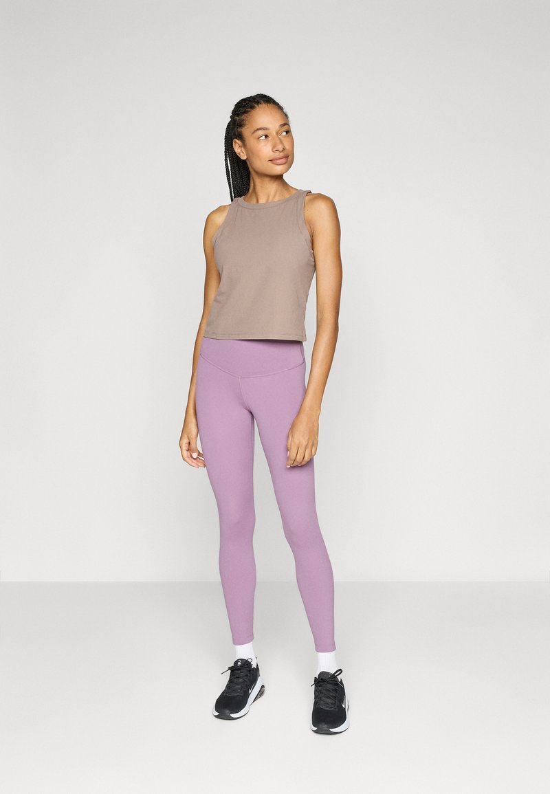 Mujer con el cabello trenzado que lleva una blusa sin mangas beige, leggings lavanda, calcetines blancos y zapatillas negras, de pie y mirando hacia un lado.